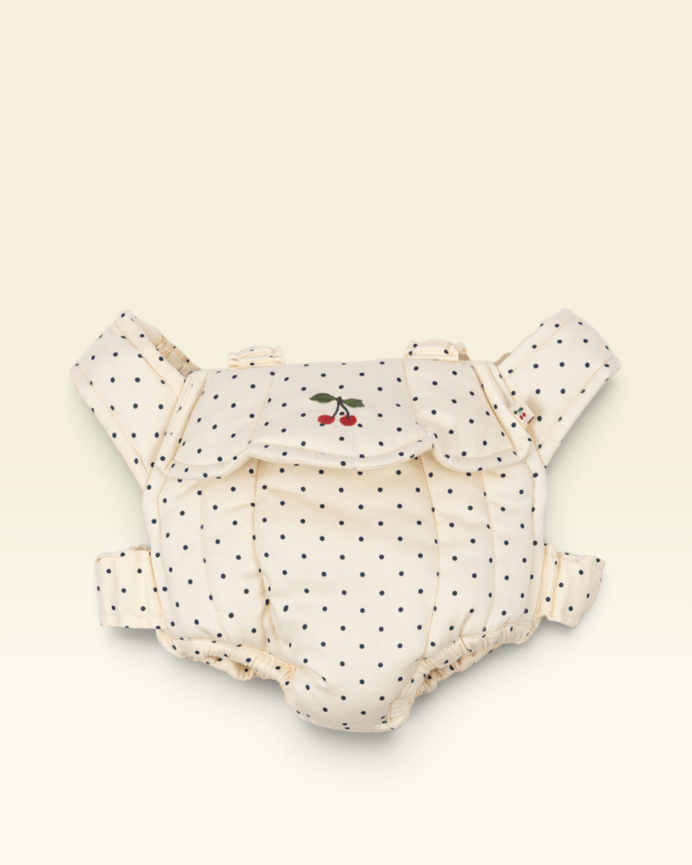 A cream-colored baby carrier with black polka dots and a small embroidered cherry motif on the front flap, displayed against a solid cream background.