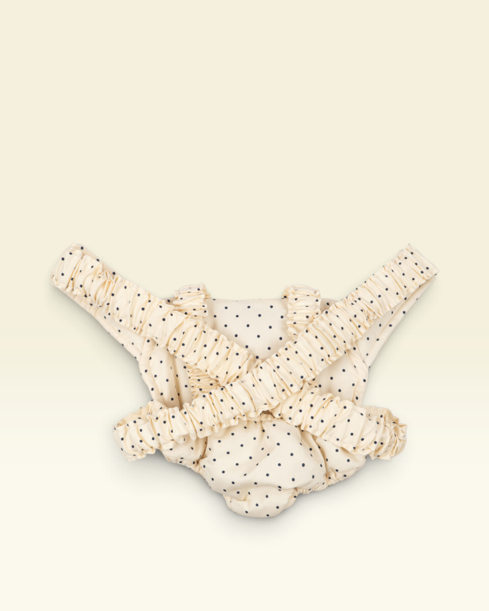 A cream-colored baby harness with black polka dots and ruffled straps, laid flat on a light cream background.