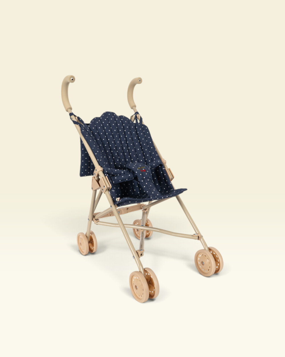A miniature toy baby stroller with a navy blue fabric seat decorated with small white polka dots, supported by a light beige metal frame with wooden-look wheels and handles, set against a solid cream background.