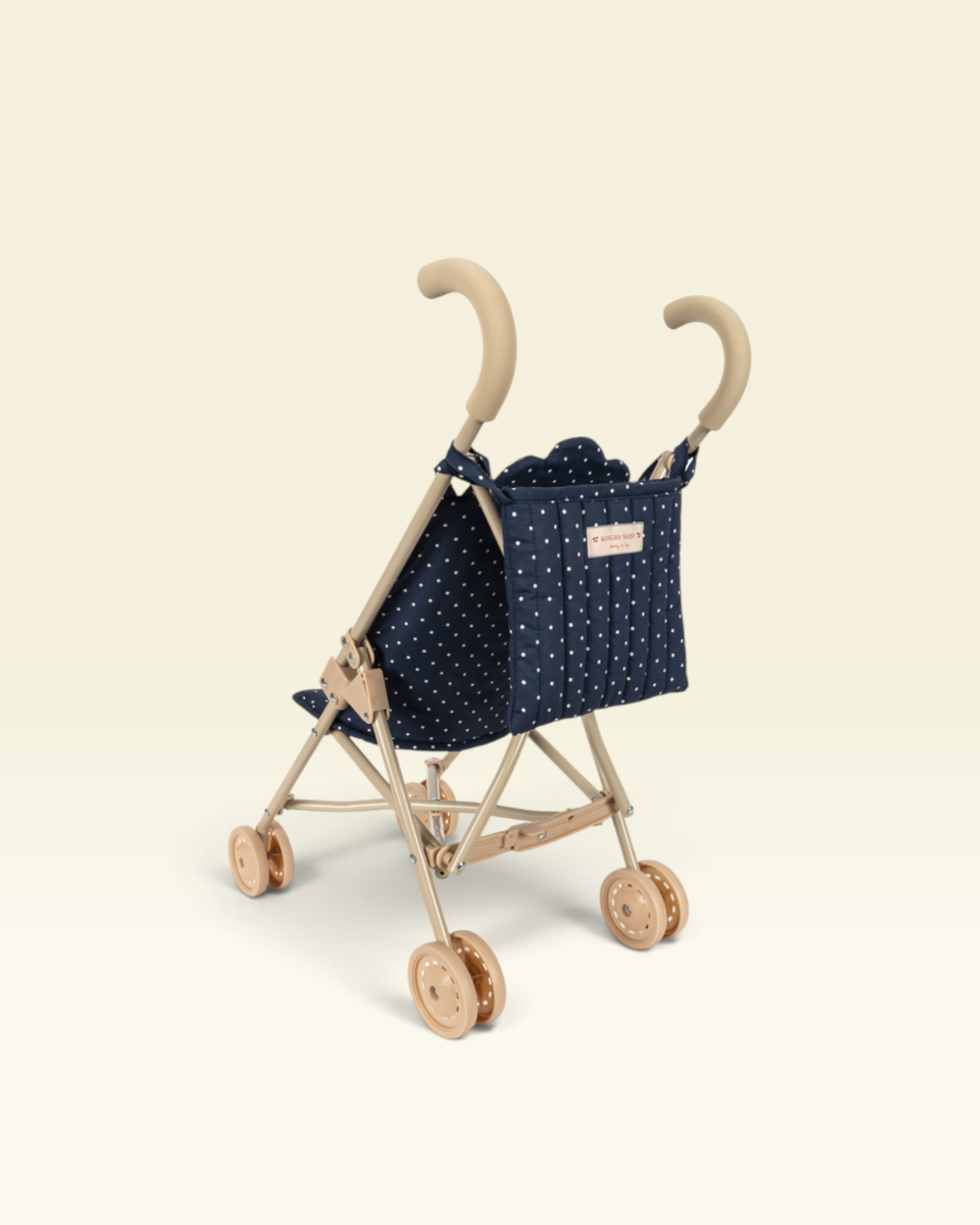 A compact stroller with a natural wood frame, beige handles, and a navy blue fabric seat cover decorated with small white polka dots. The stroller is photographed from a high-angle perspective against a solid, warm off-white background.