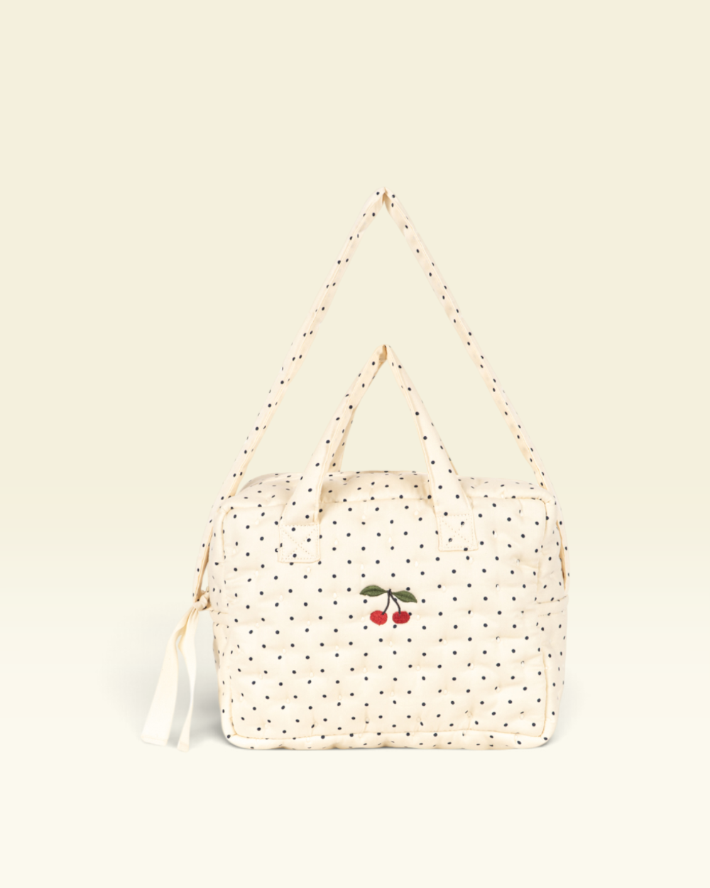 A cream-colored quilted bag with black polka dots and a small embroidered cherry motif on the front, featuring a matching shoulder strap and top handles, set against a solid cream background.