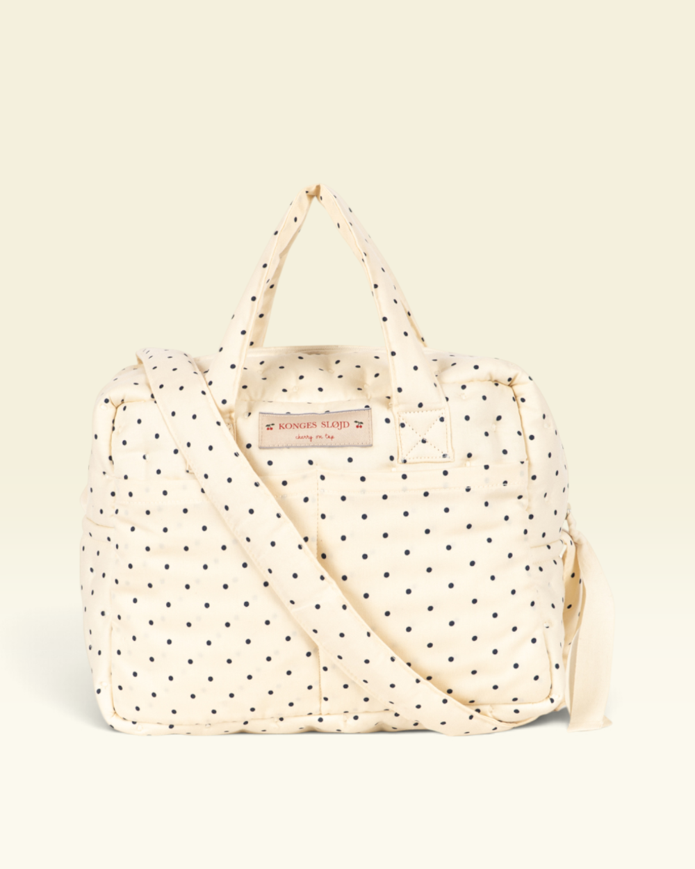 An off-white quilted duffel bag with black polka dots, featuring dual top handles, a detachable shoulder strap.