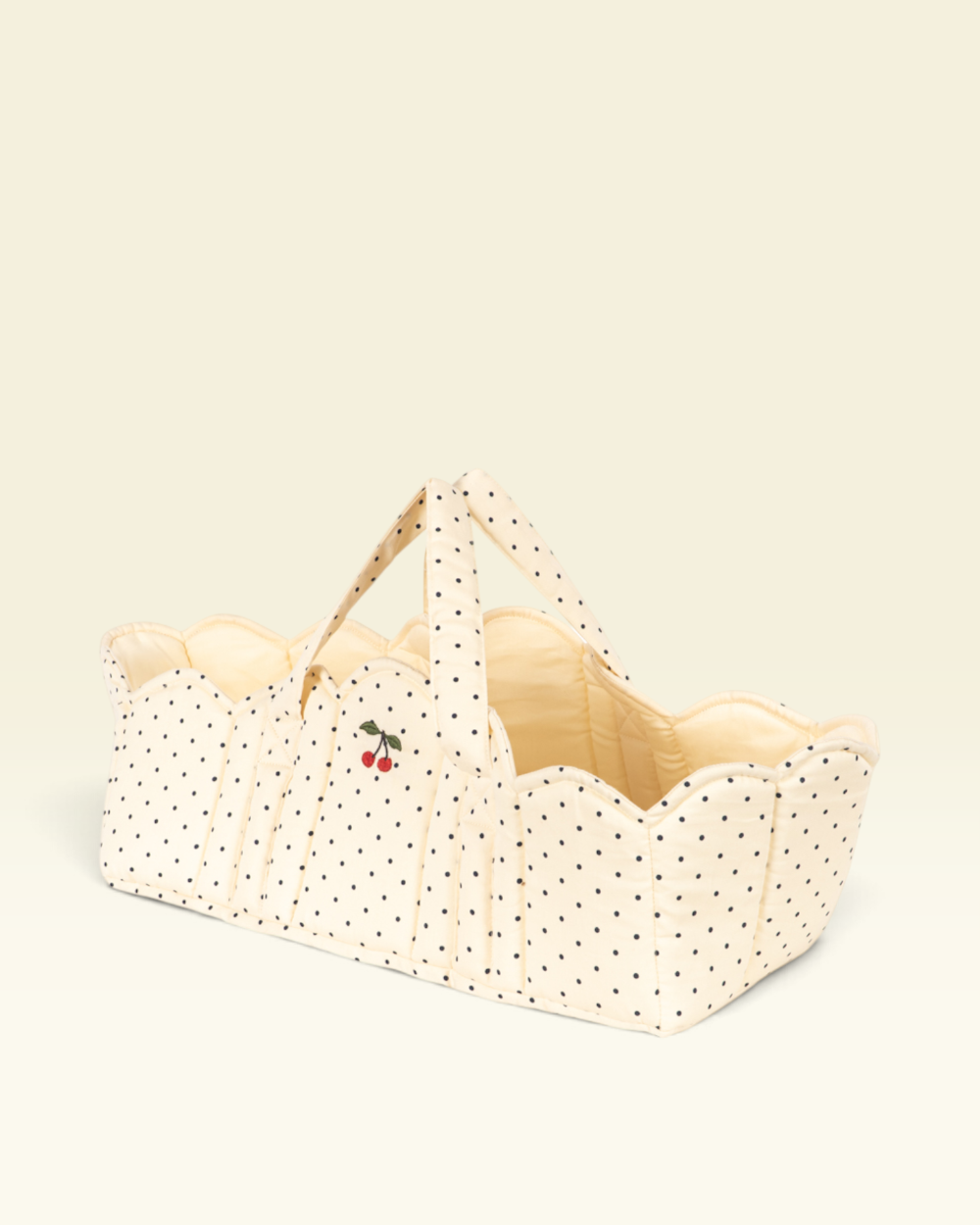 A cream-colored fabric bassinet or basket with black polka dots, featuring a small cherry embroidery on the front and matching fabric handles, set against a solid cream background.