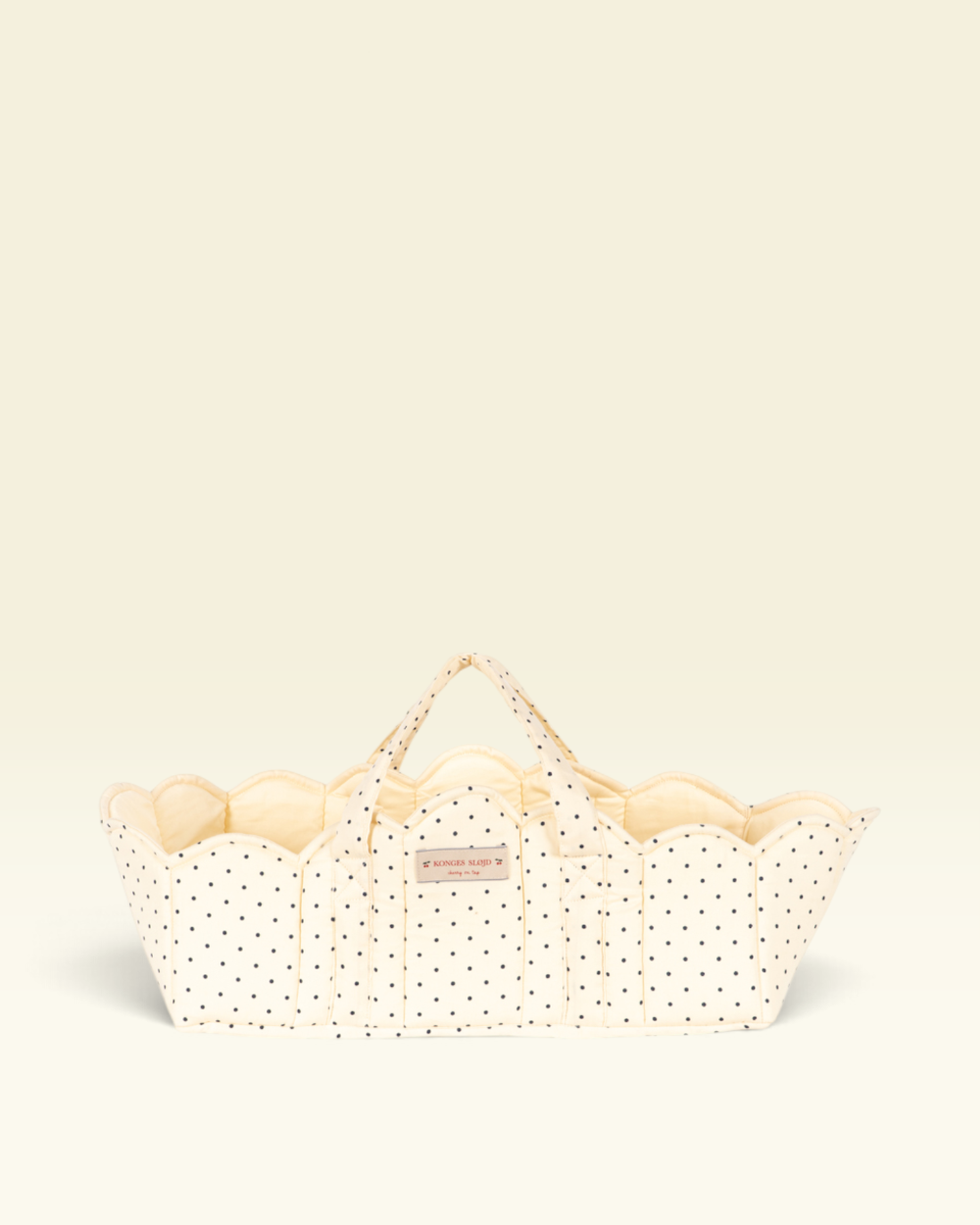 A cream-colored fabric basket with black polka dots and scalloped edges, featuring two top handles and a small pink brand label, set against a solid cream background.