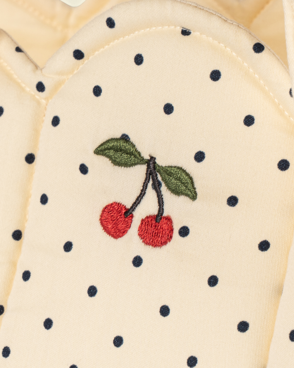Close-up of a cream-colored fabric garment with black polka dots and an embroidered cherry motif in red, green, and black.
