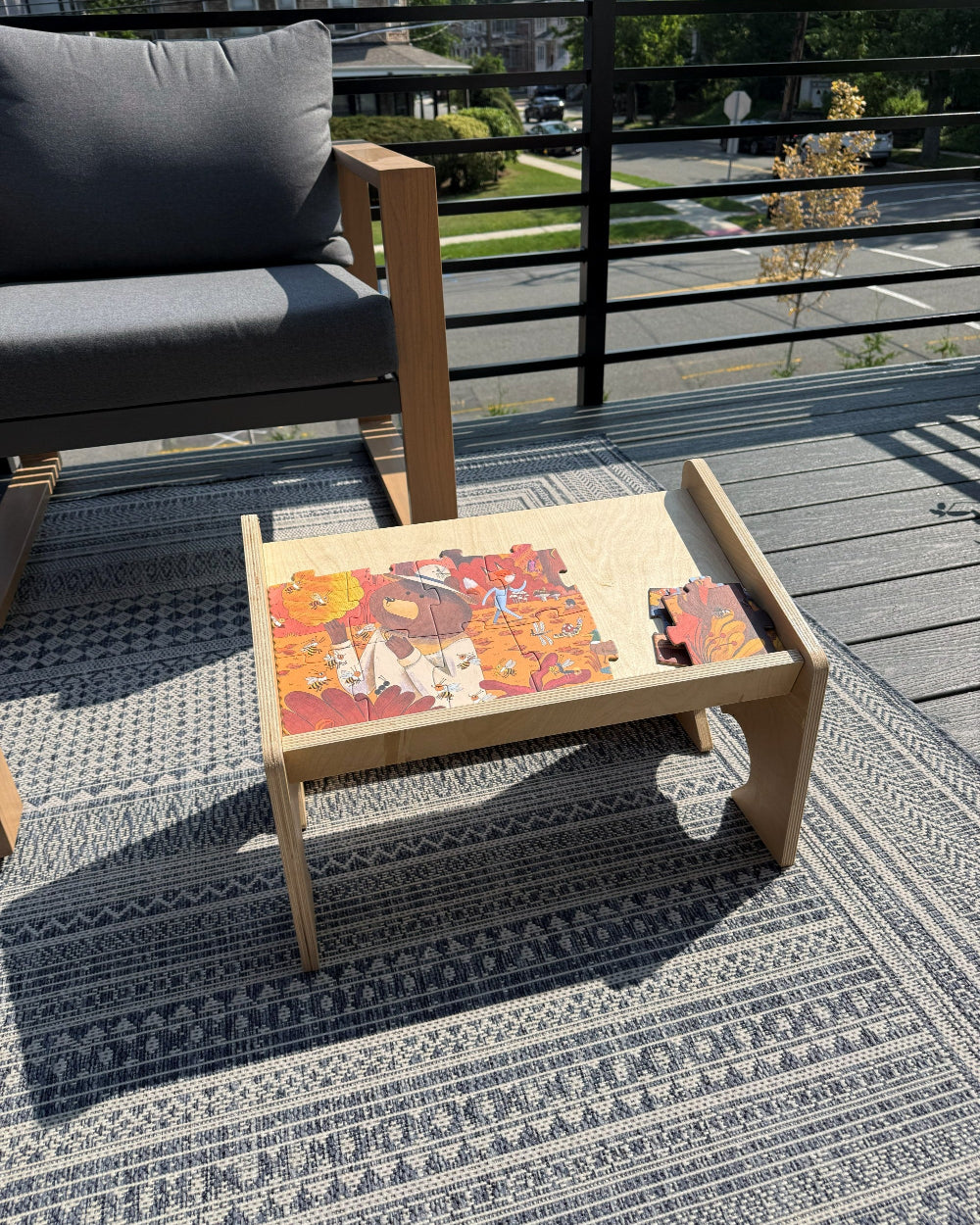 A light natural wood low coffee table with a slanted tray top, styled on a patterned outdoor rug on a wooden deck. The tray contains a colorful jigsaw puzzle and a small red toy.