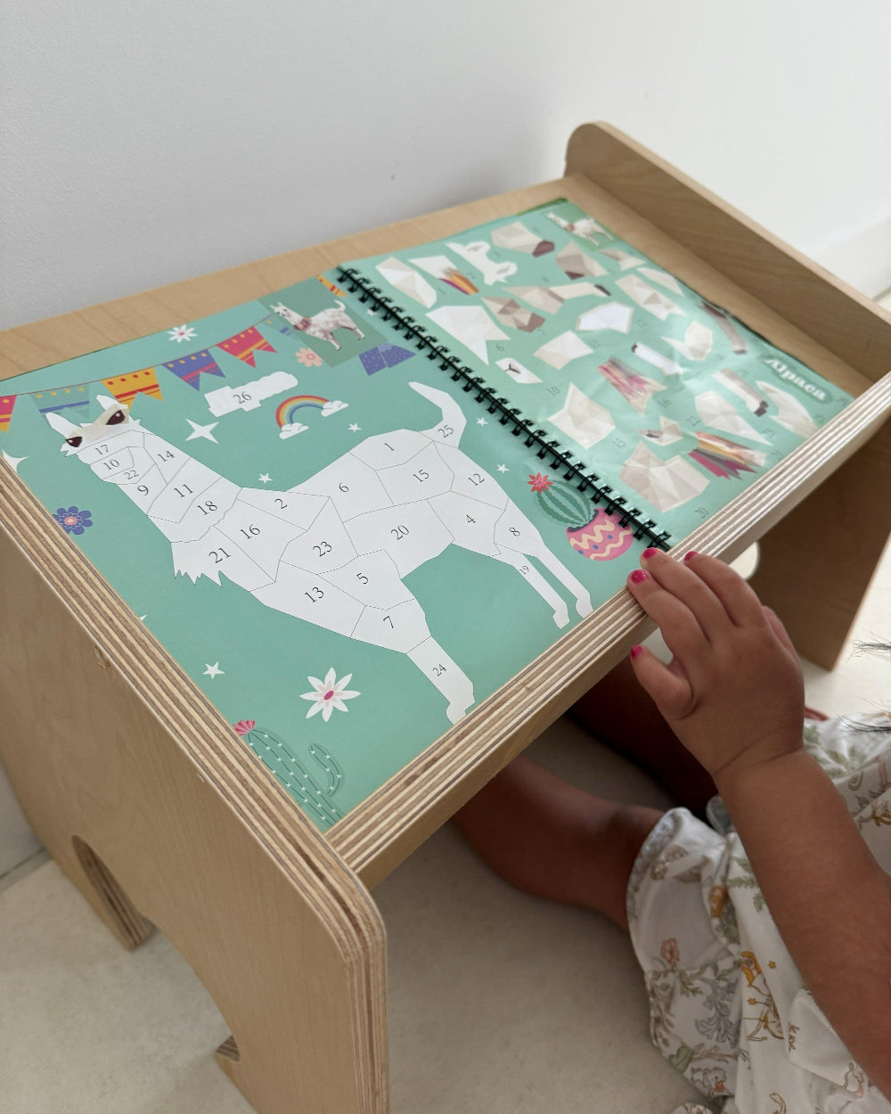 A child's hand is shown interacting with a wooden desk featuring a colorful llama puzzle board. The desk has a light natural wood finish and a mint green puzzle cover decorated with cartoon llamas, stars, and flags.
