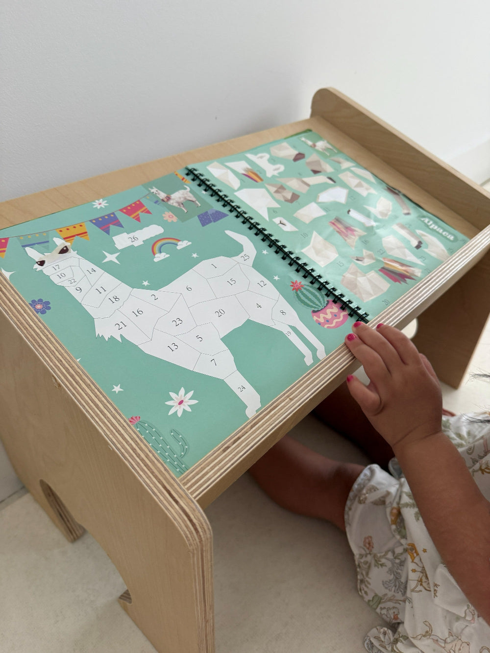A child's hand is shown interacting with a wooden desk featuring a colorful llama puzzle board. The desk has a light natural wood finish and a mint green puzzle cover decorated with cartoon llamas, stars, and flags.
