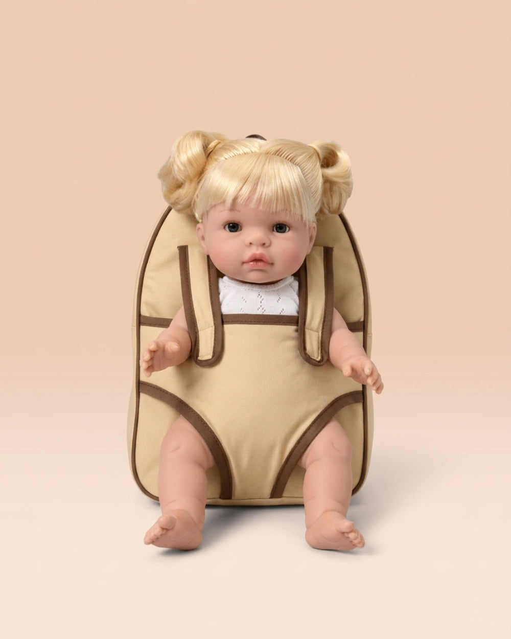 A blonde doll with pigtails sits inside a beige baby carrier or backpack-style seat with brown trim, photographed against a soft peach studio background.