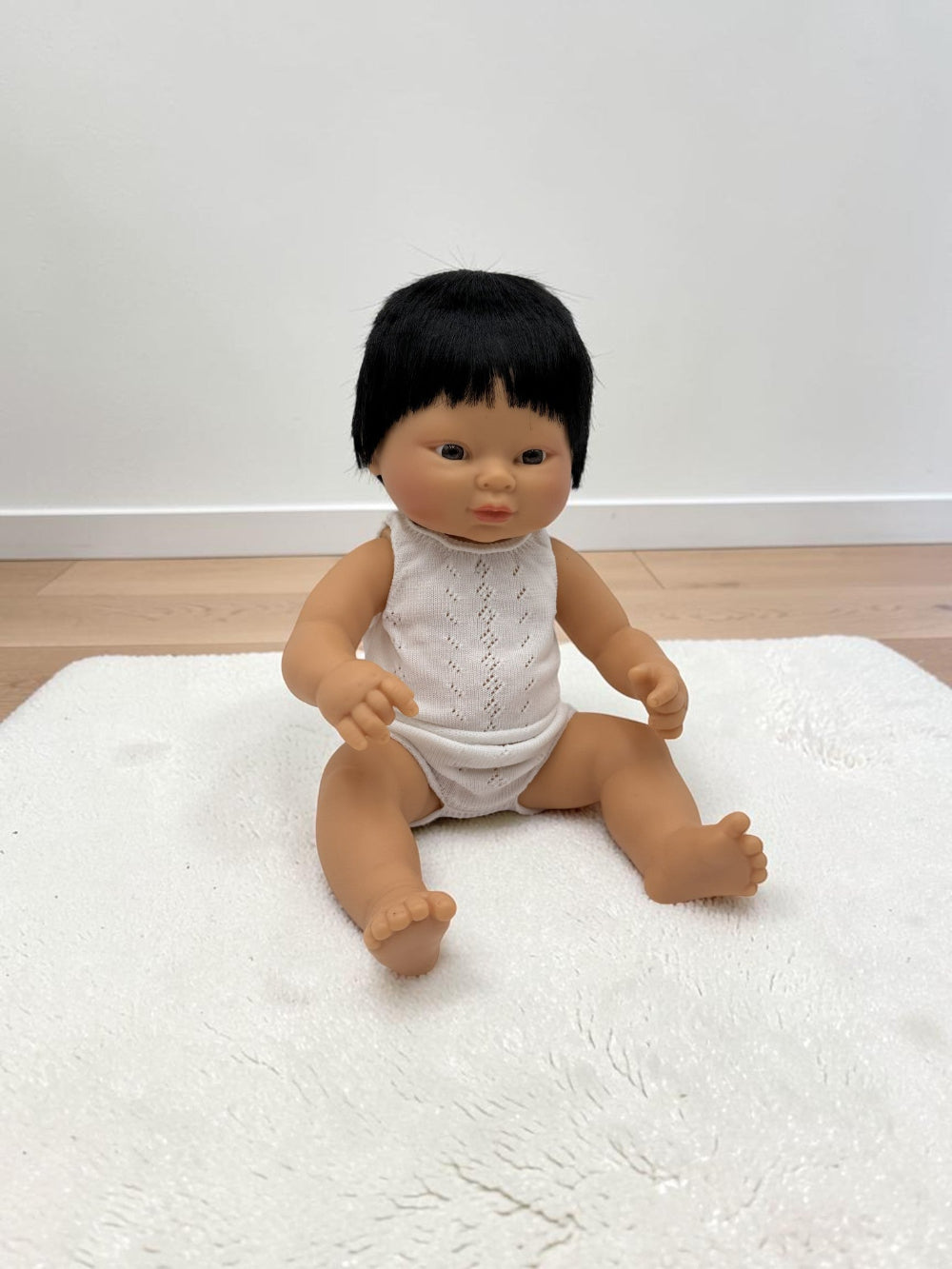 A realistic baby doll with black hair wearing a white sleeveless knit bodysuit, sitting on a white textured rug in a room with light wood flooring and a white wall.