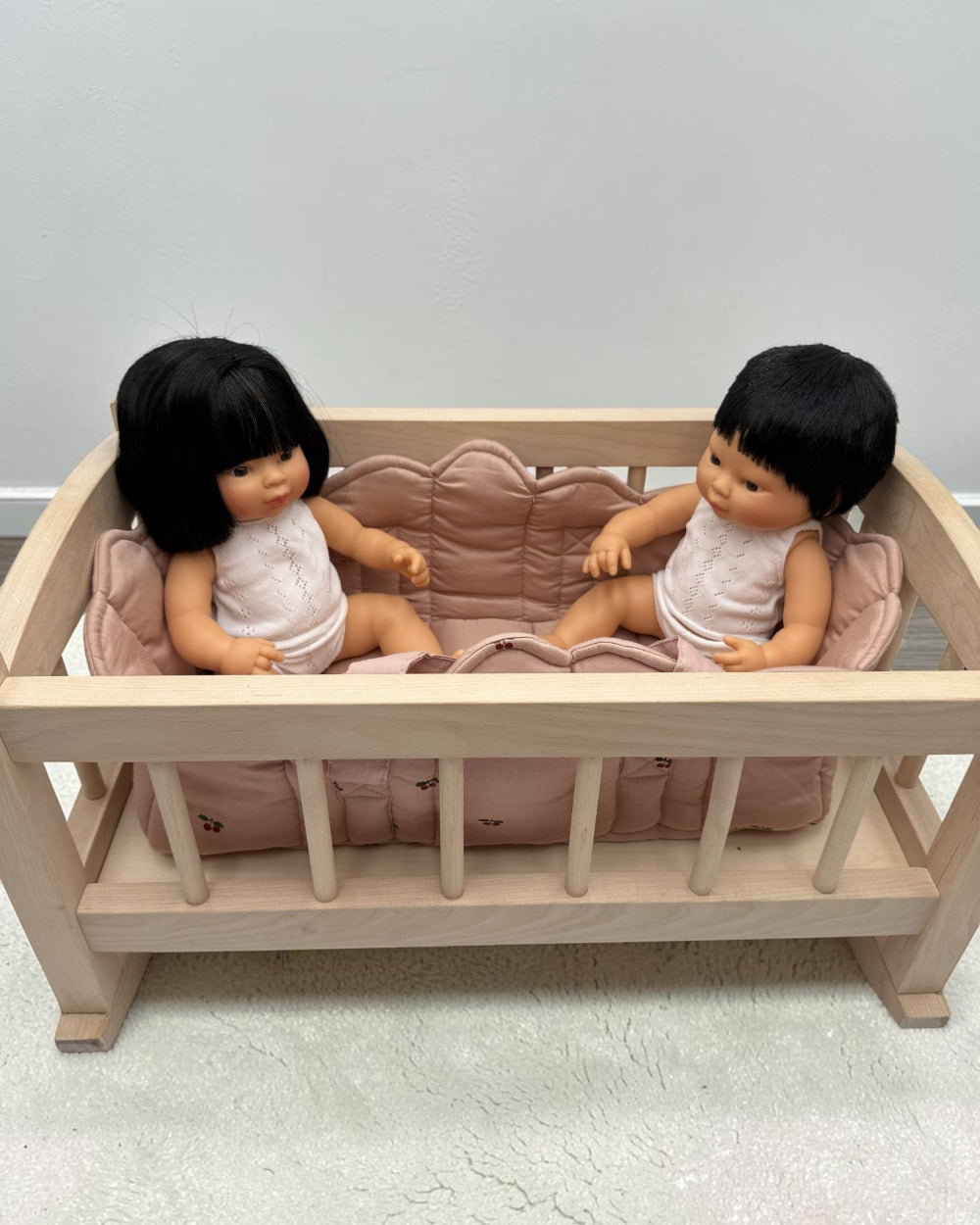 A light natural wood doll bed with a pink cushion, containing two small dolls with black hair and white outfits, set against a plain light gray wall and textured floor.