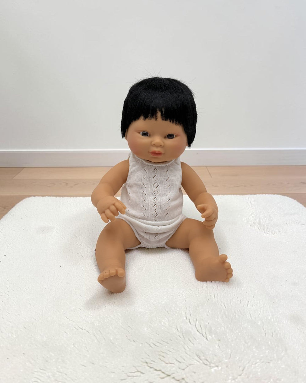 A baby doll with black hair wearing a white sleeveless top and white diaper cover, sitting on a white textured rug against a plain white wall and light wood floor.