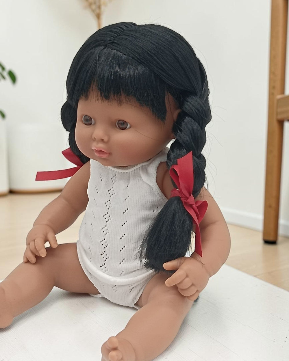 Doll with braided black hair and red ribbons wearing a white outfit on a light surface.