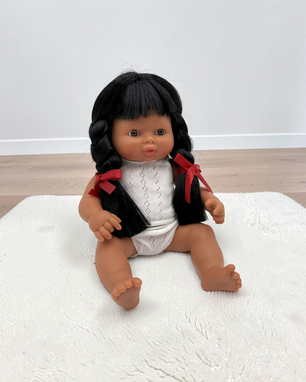 A baby doll with black braided hair and red ribbon accents, wearing a white knit top and white diaper, sitting on a white textured rug.