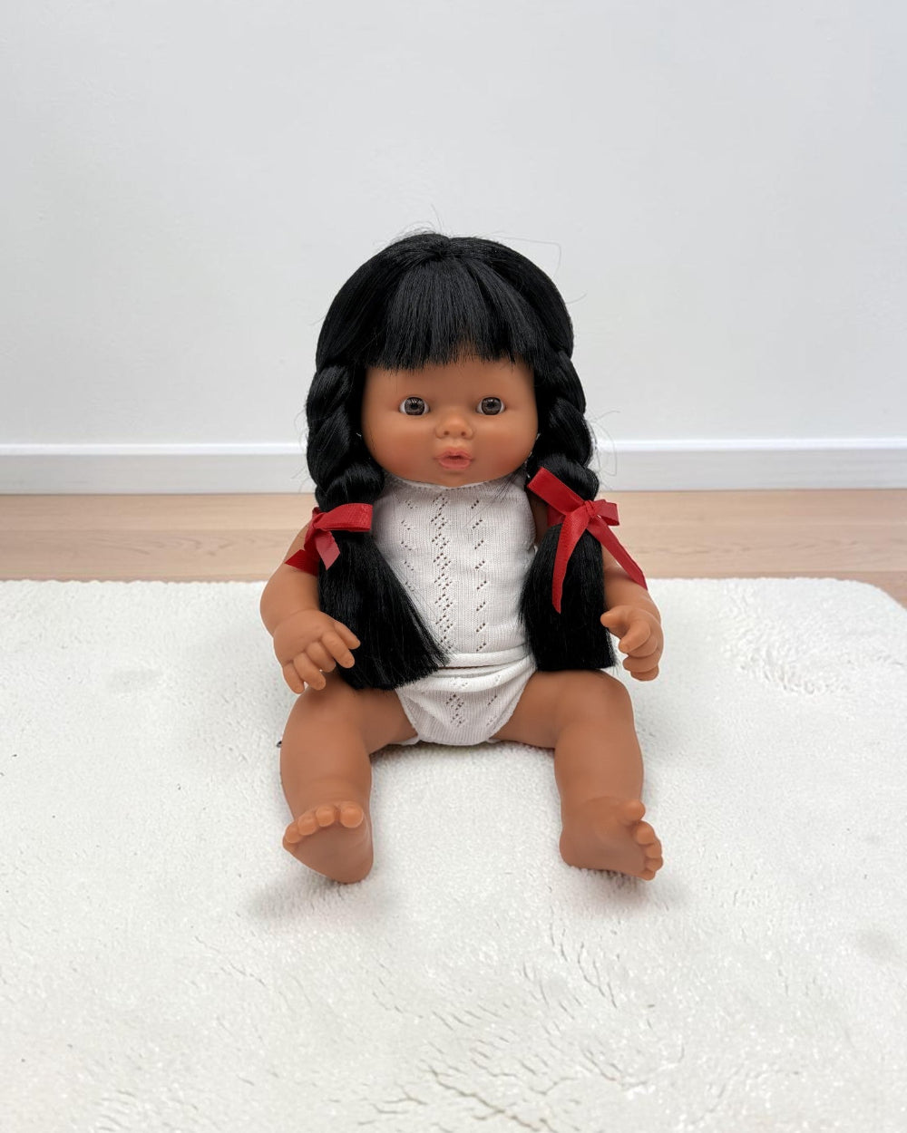 A realistic baby doll with dark brown skin, black braided hair, and a white knit top with red ribbon accents, sitting on a white textured rug.