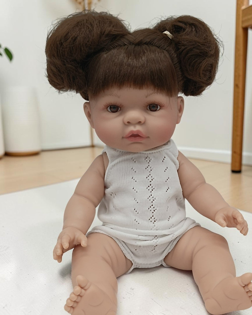 Realistic baby doll with brown hair styled in buns wearing a light gray romper on a white surface.
