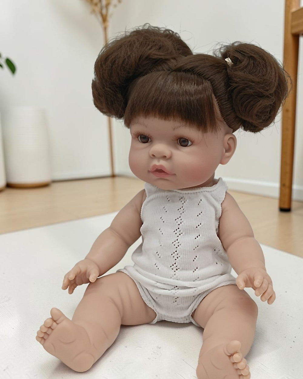 Baby doll with brown hair styled in buns wearing a white sleeveless outfit on a light surface.