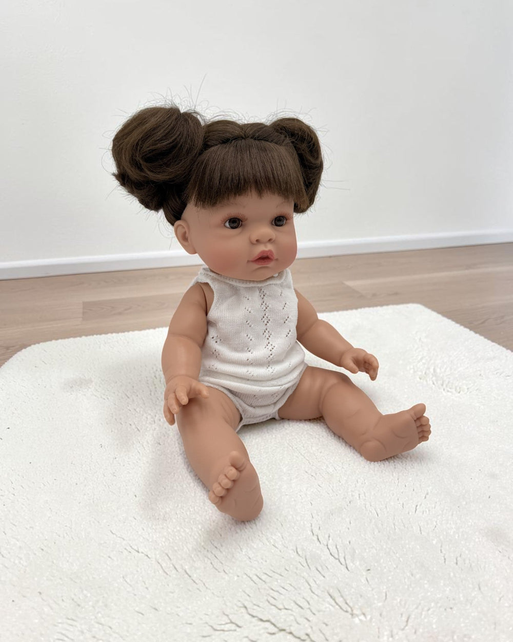 A realistic baby doll with brown hair styled in two buns sits on a white textured rug in a room with light wood flooring and a white wall.