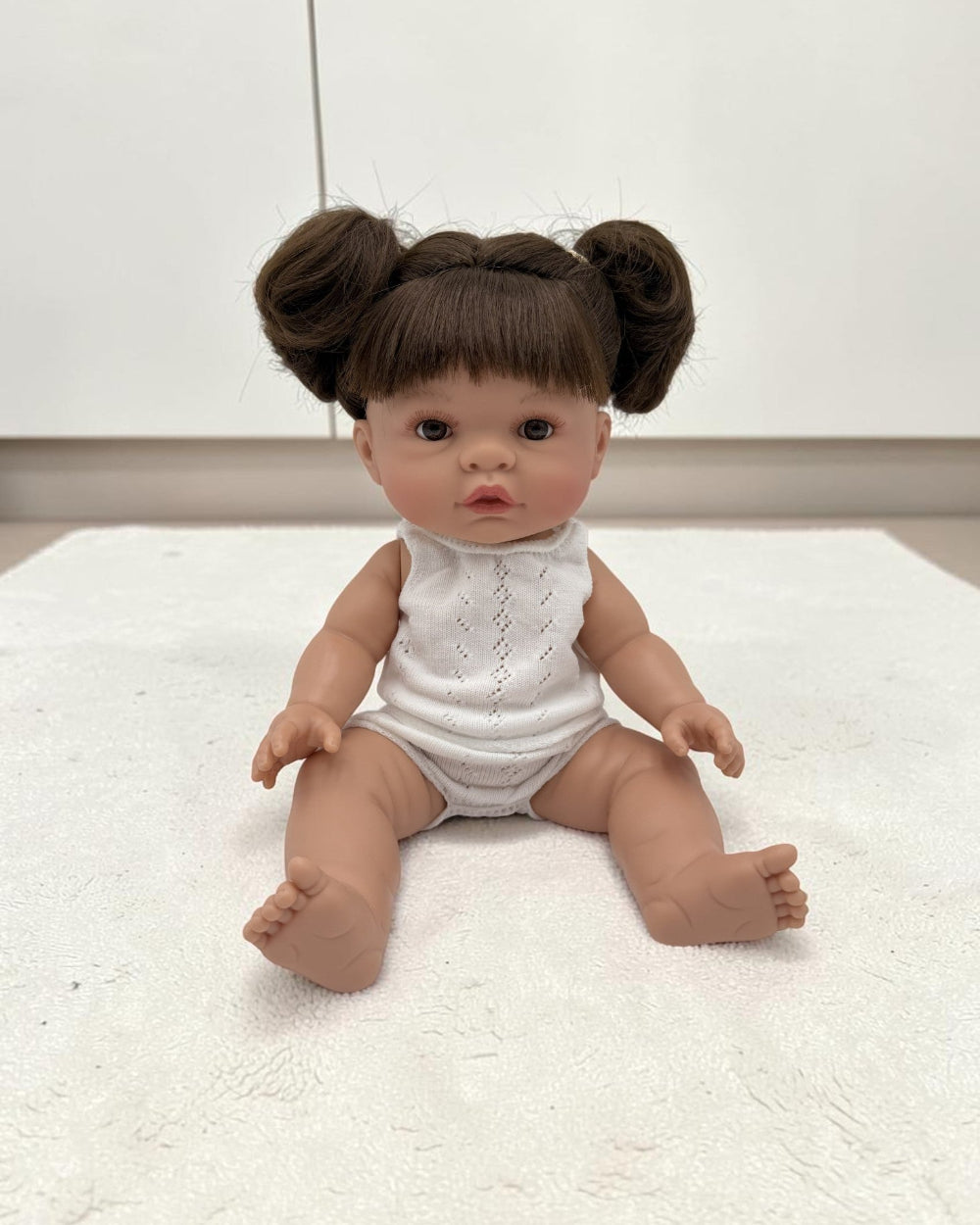 A realistic baby doll with brown hair styled in two buns, wearing a white sleeveless bodysuit, sitting on a white textured rug.