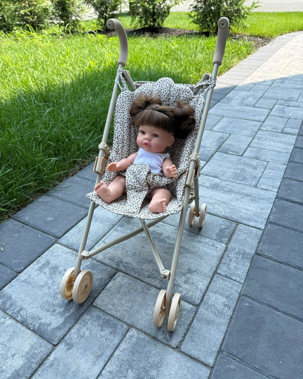 A vintage-style doll stroller with a beige frame and patterned fabric seat, containing a baby doll with brown hair, placed on a gray stone pathway with green grass in the background.