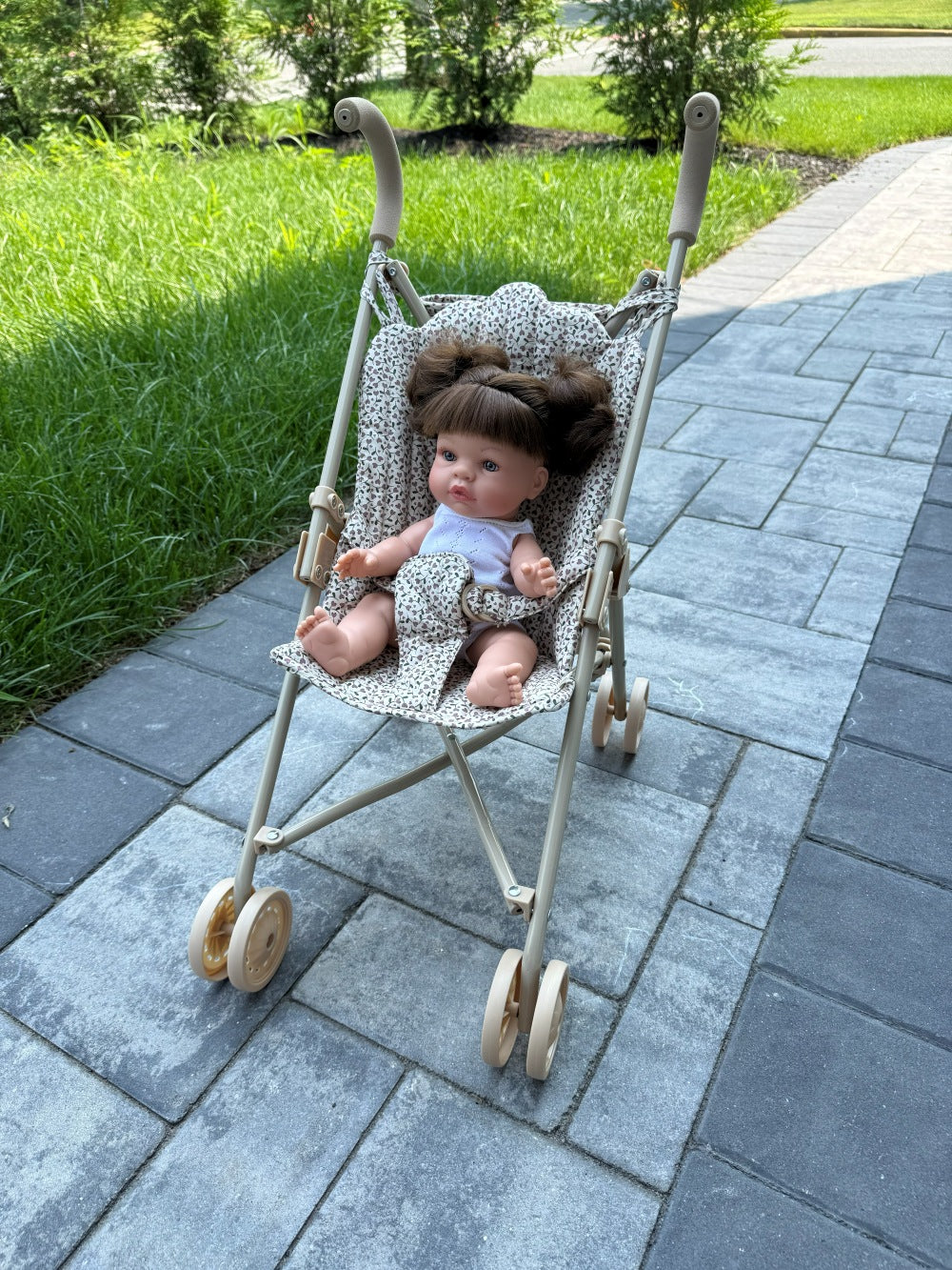 A vintage-style doll stroller with a beige frame and patterned fabric seat, containing a baby doll with brown hair, placed on a gray stone pathway with green grass in the background.