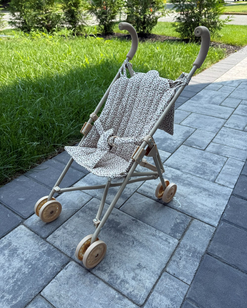 A stroller with a beige frame and a patterned cream and brown fabric seat cover, parked on a gray paved pathway with green grass and trees in the background.