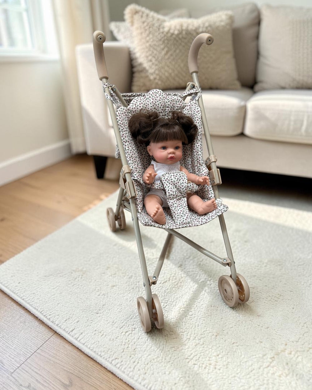 A miniature doll stroller with a gray and white patterned fabric seat, shown on a cream rug in a bright living room with a doll sitting inside.