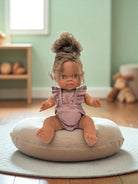 Doll sitting on a cushion in a room with a green wall and wooden floor.