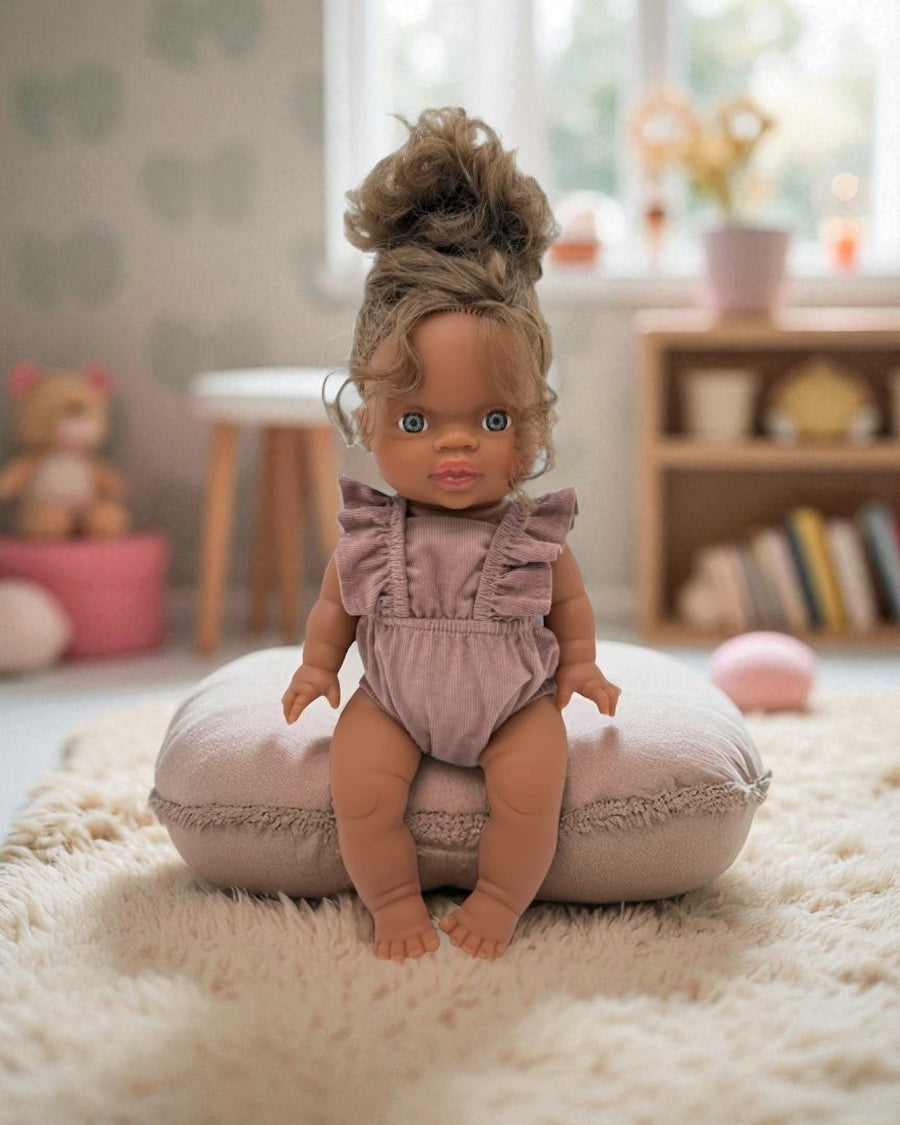 Doll in a chestnut outfit sitting on a cushion in a room with toys and books.