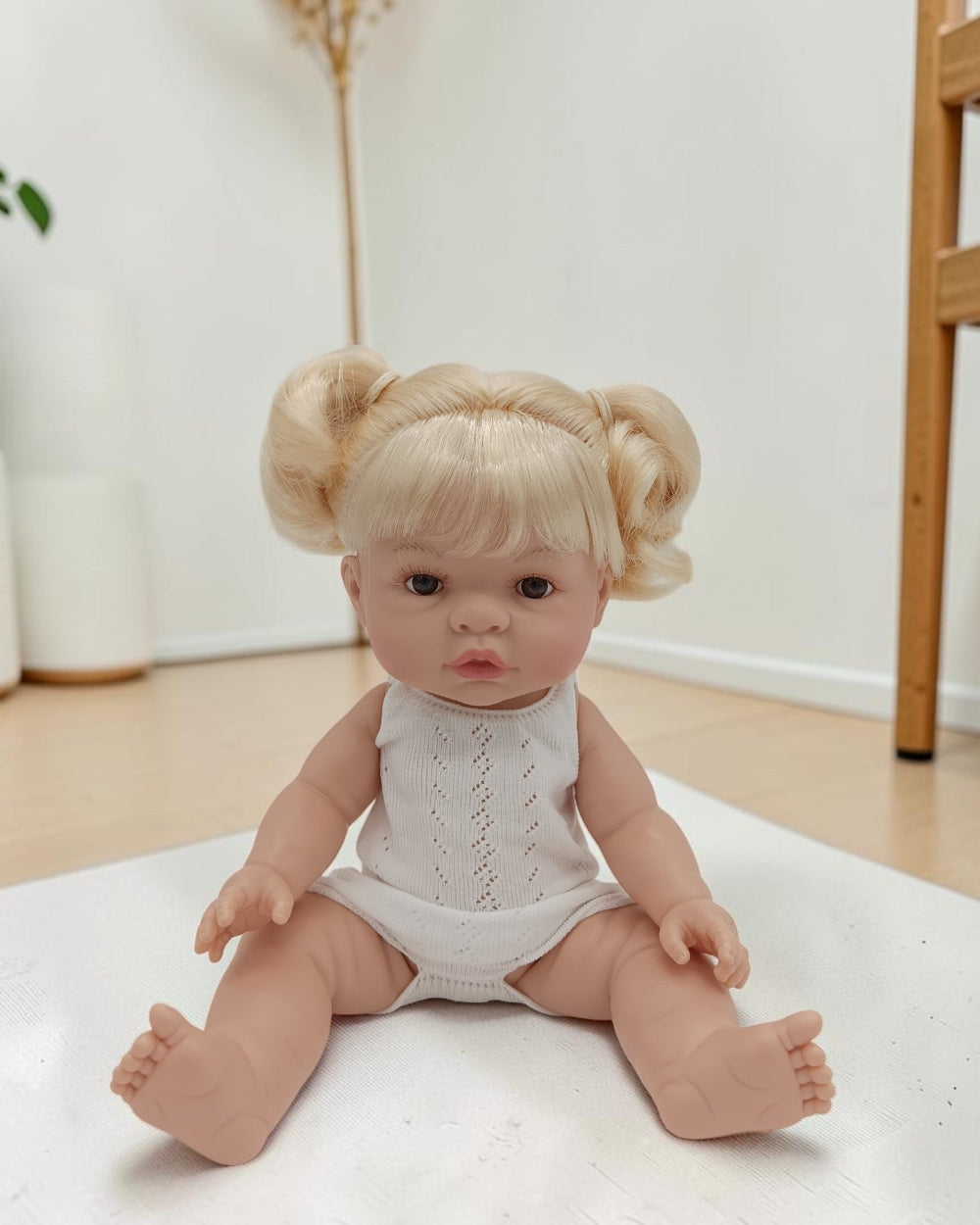 Baby doll with blonde hair styled in pigtails wearing a white onesie on a light surface.