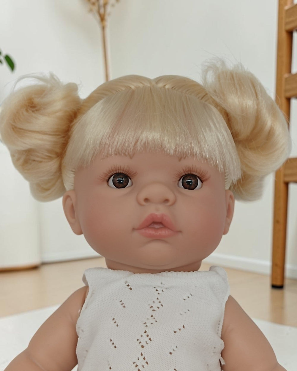 Baby doll with blonde hair styled in pigtails wearing a light gray dress indoors.