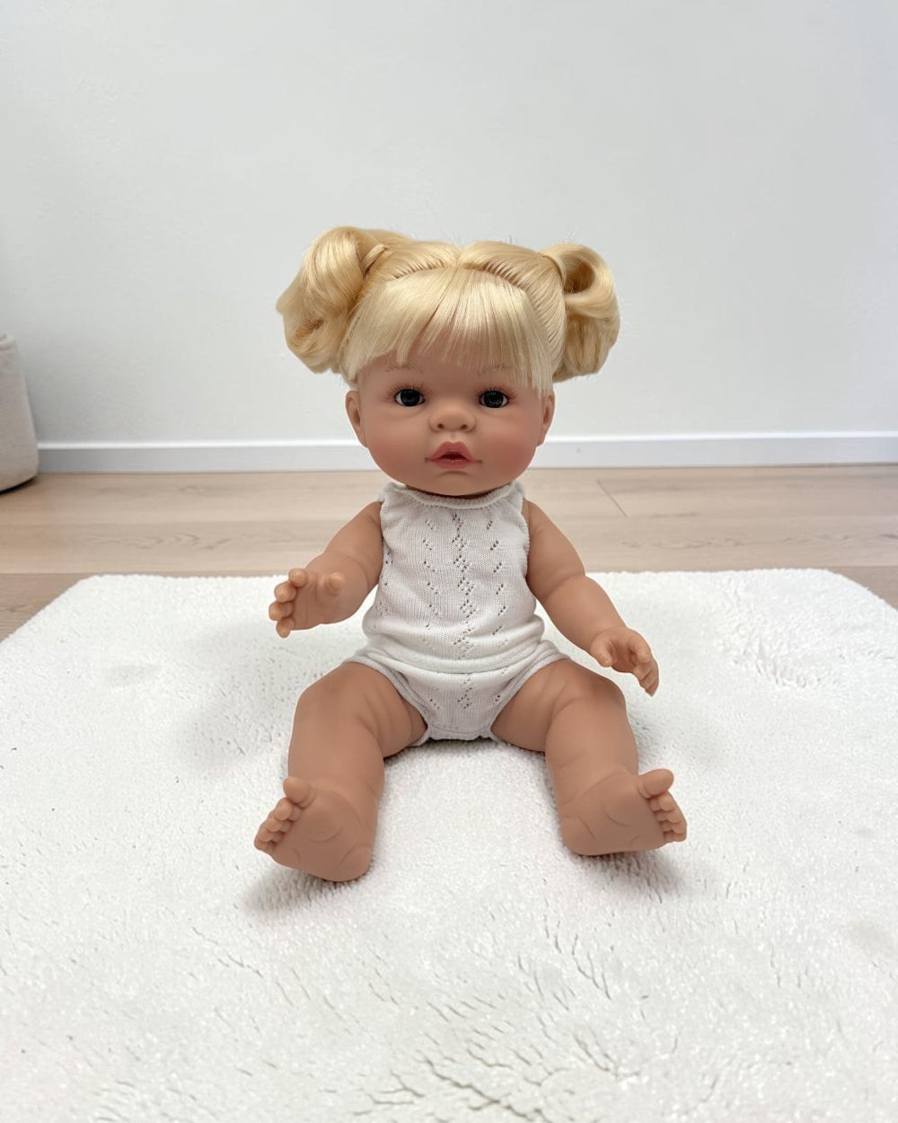 A realistic baby doll with blonde hair in two buns sits on a white textured rug in a room with light wood flooring and a white wall.