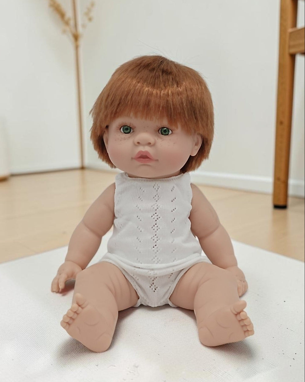 Baby doll with red hair wearing a white lace romper on a light surface.
