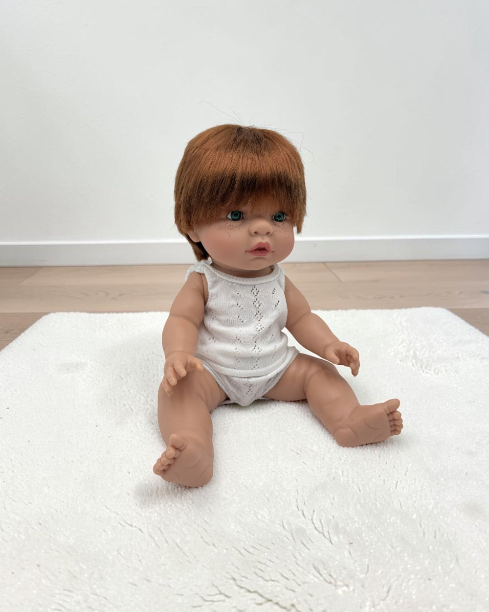 A realistic baby doll with brown hair sits on a white textured rug in a room with light wood flooring and a white wall.