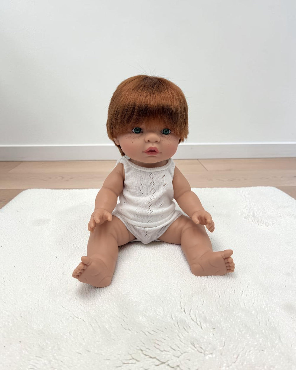 A realistic baby doll with short brown hair sits on a white textured rug against a plain white wall and light wood floor.