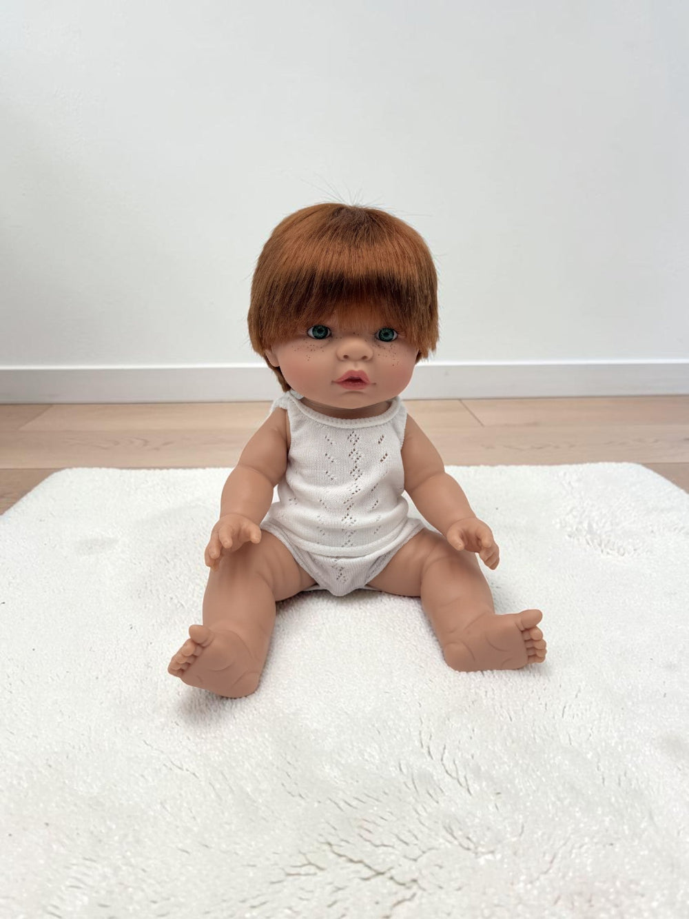 A realistic baby doll with short brown hair sits on a white textured rug against a plain white wall and light wood floor.