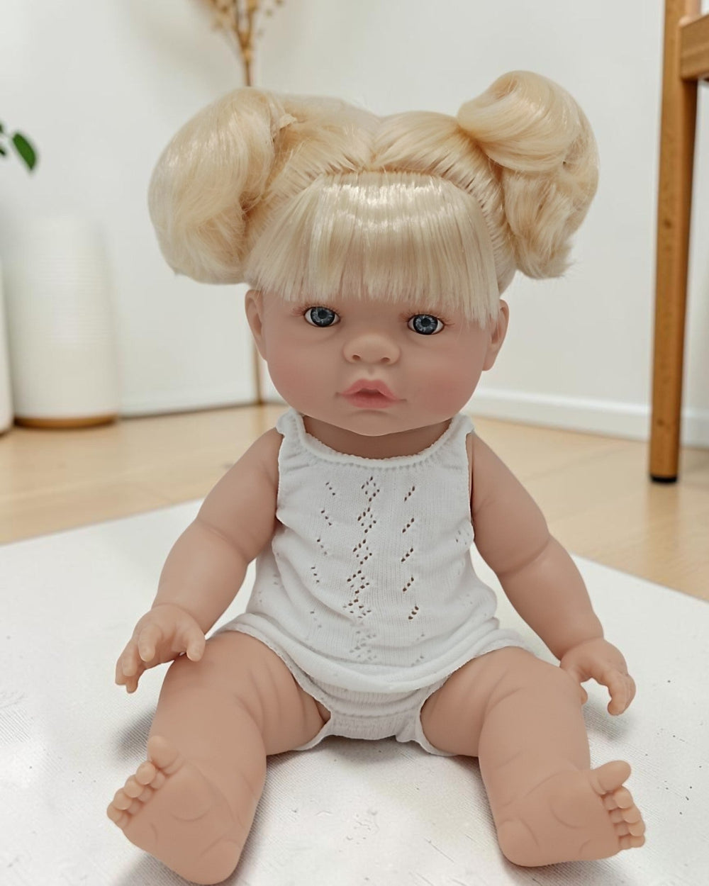 Realistic baby doll with blonde hair styled in buns, wearing a white outfit, on a light-colored surface.