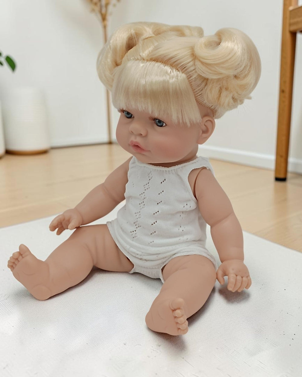 Baby doll with blonde hair styled in buns, wearing a white outfit, on a light-colored surface.