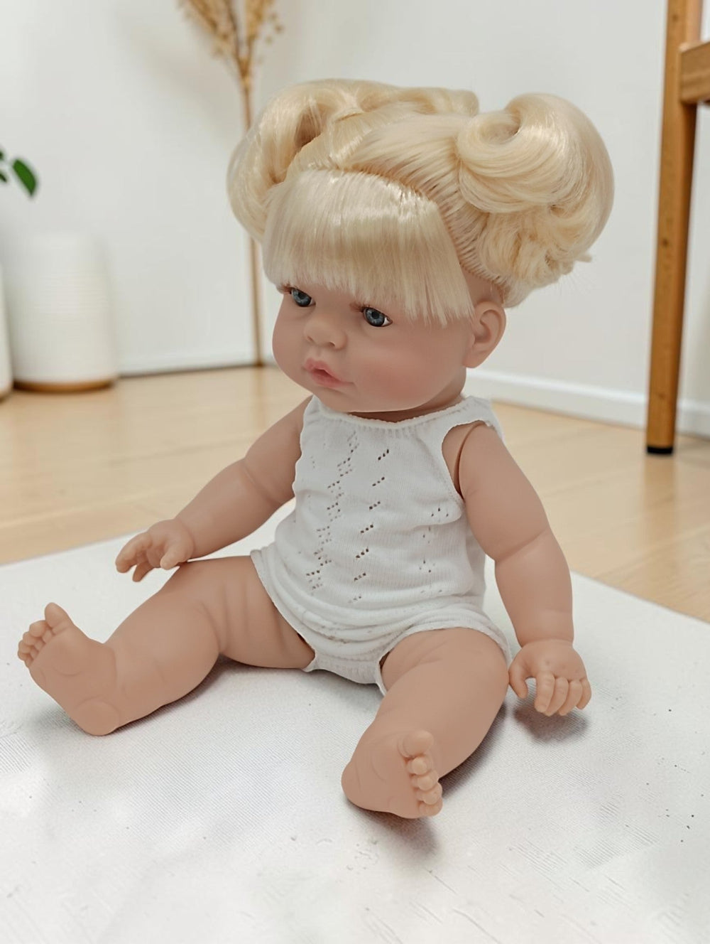 Baby doll with blonde hair styled in buns, wearing a white outfit, on a light-colored surface.