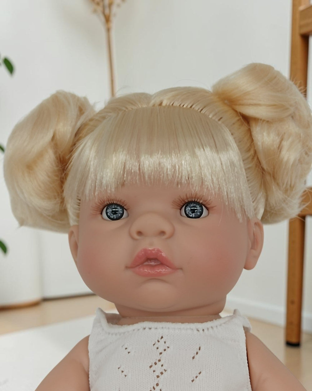 Doll with blonde hair styled in pigtails and blue eyes wearing a white top.