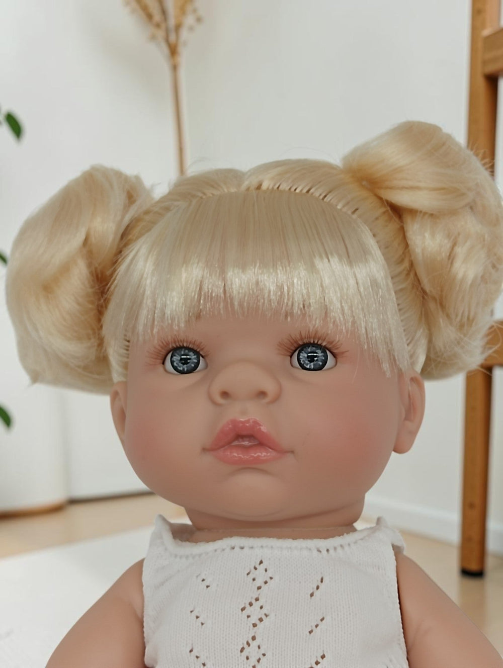 Doll with blonde hair styled in pigtails and blue eyes wearing a white top.