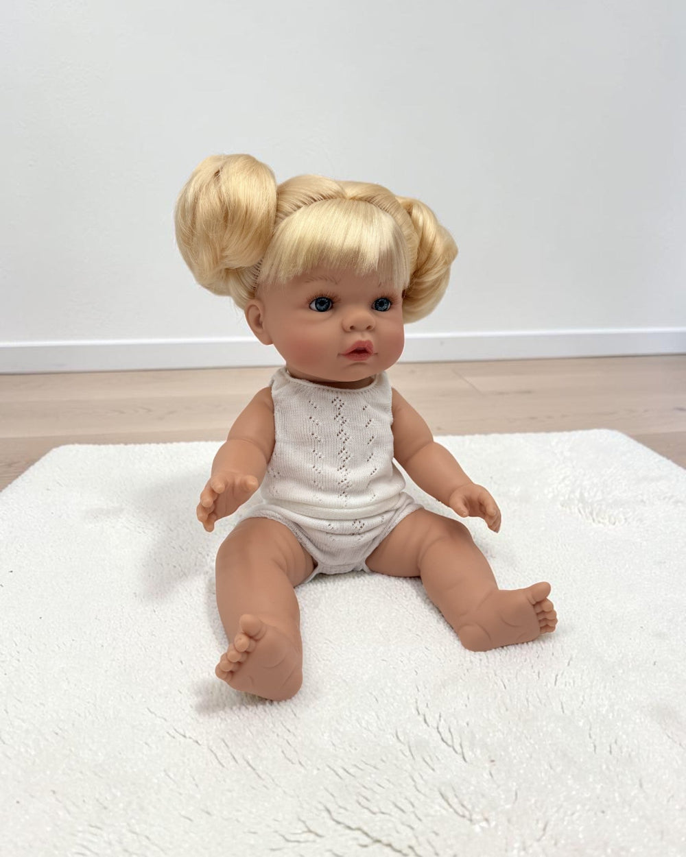 A baby doll with blonde hair styled in two buns sits on a white textured rug against a plain white wall and light wood floor.
