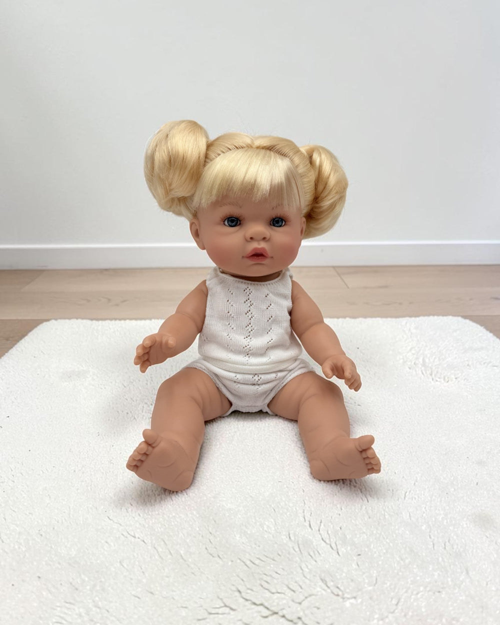 A realistic baby doll with blonde pigtails sits on a white textured rug in front of a plain white wall and light wood floor. The doll is wearing a white sleeveless knit romper.