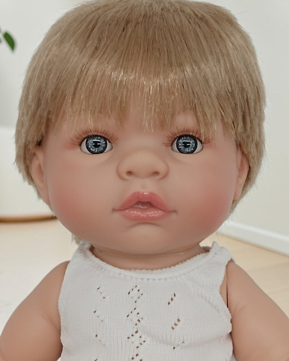Realistic baby doll with blonde hair and blue eyes wearing a white sleeveless top.