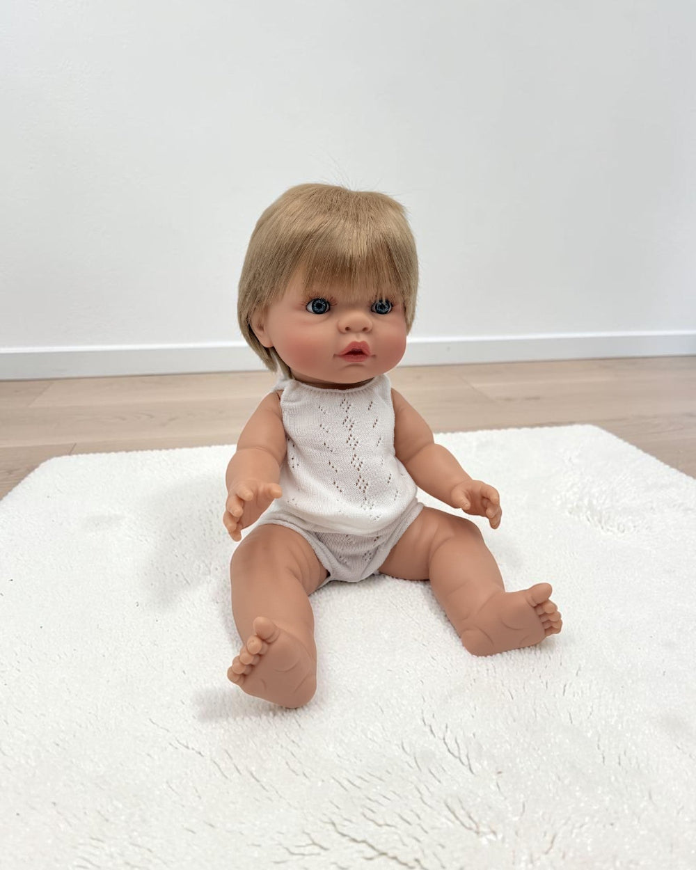 A realistic baby doll with short blonde hair and blue eyes, wearing a white sleeveless knit bodysuit, sitting on a white textured rug in a room with light wood flooring and a white wall.