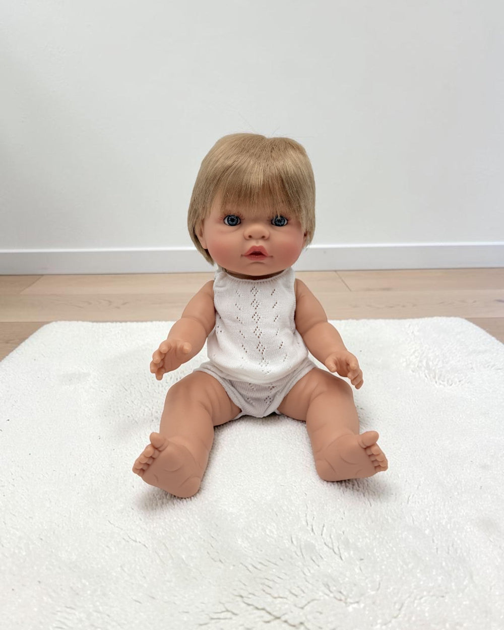 A realistic baby doll with short blonde hair and blue eyes sits on a white textured rug in a room with light wood flooring and a white wall.