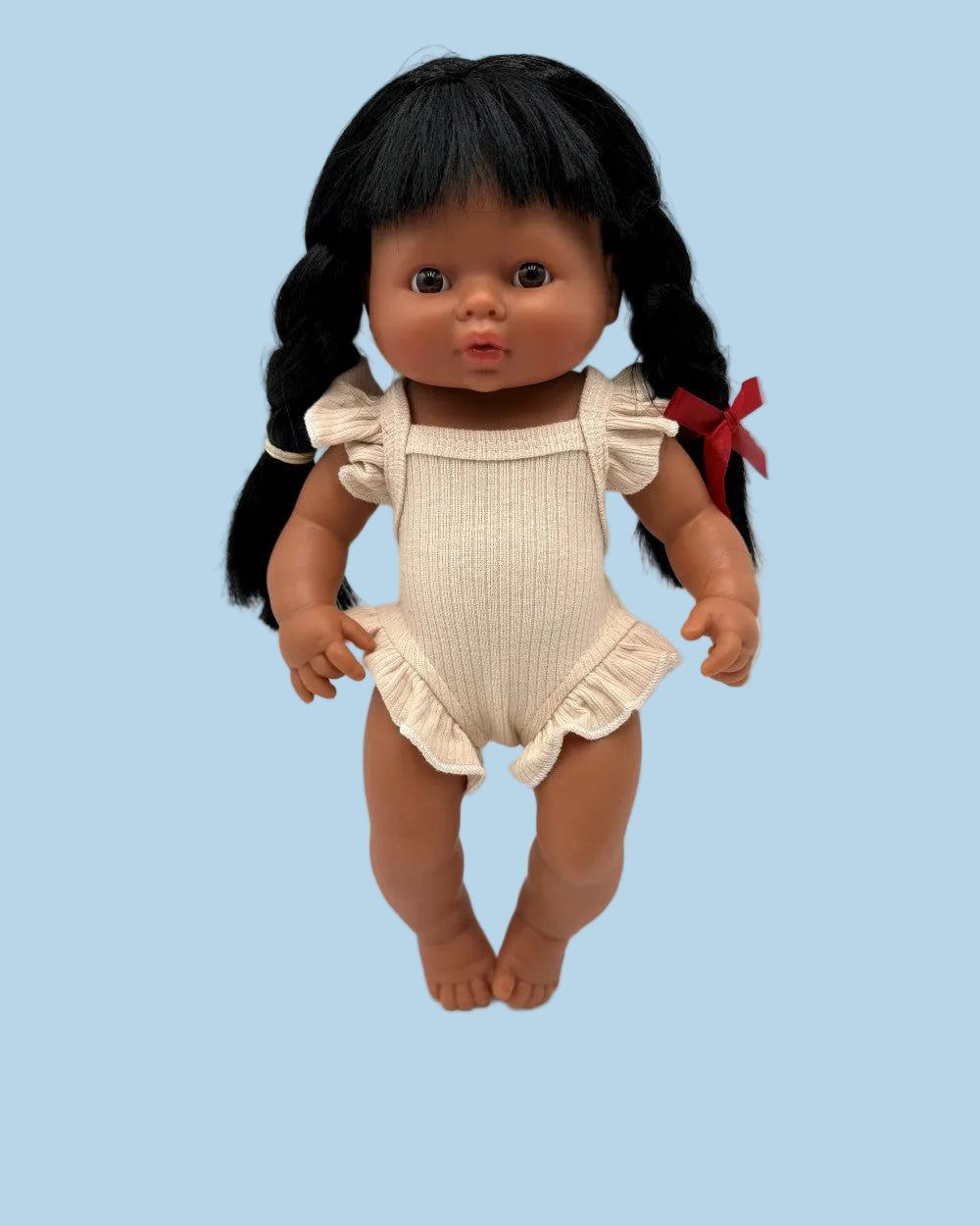 A baby doll with black hair in two braids, wearing a cream ribbed romper with ruffled sleeves and hem, and a red hair tie, standing against a plain white background.