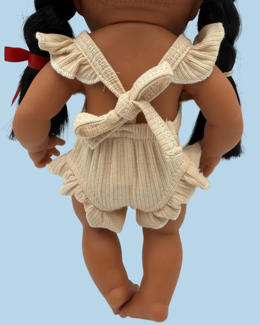 A close-up back view of a doll with dark brown skin and black pigtails, wearing a cream ribbed romper with ruffled straps and a bow tie back, photographed against a plain white background.