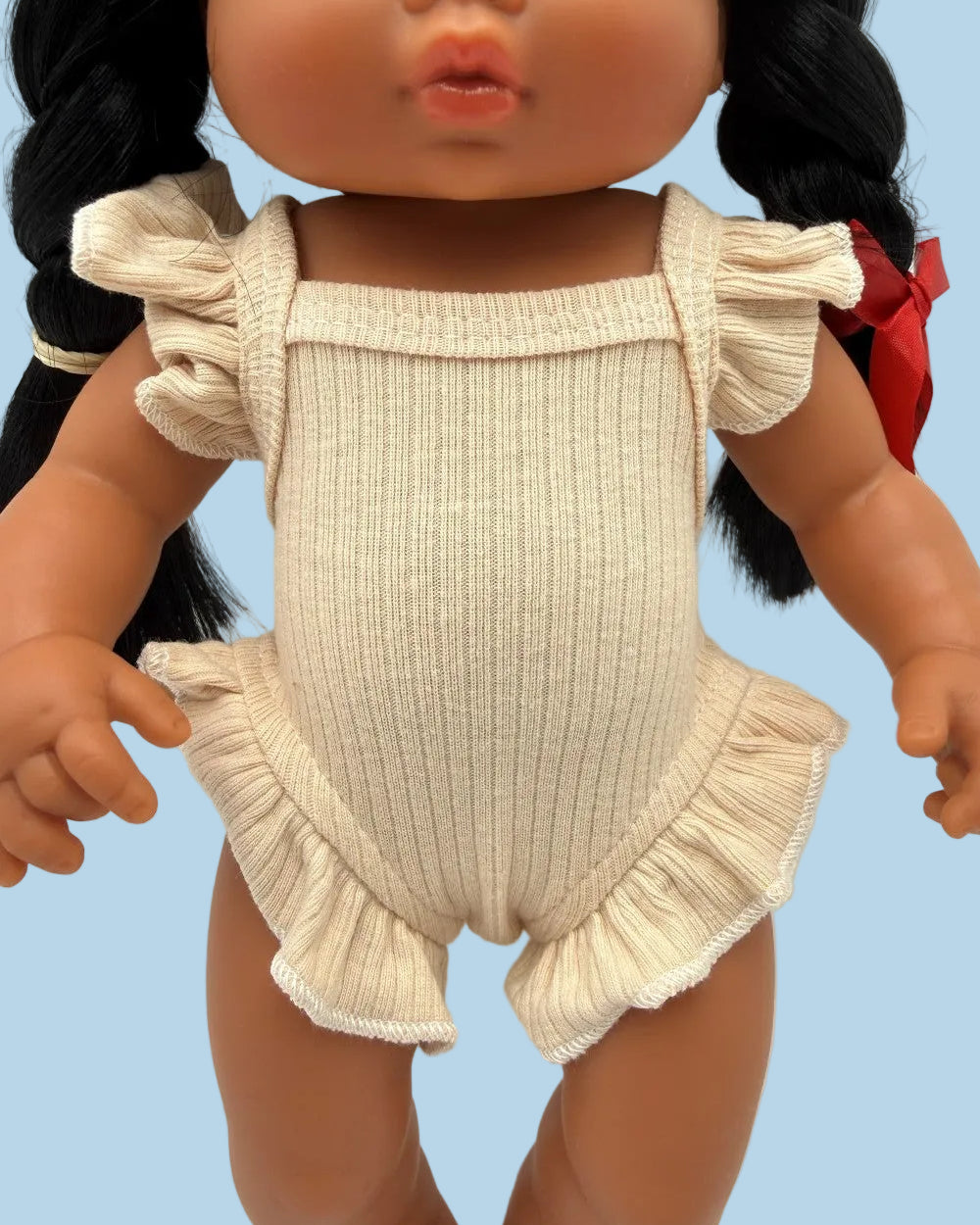 A close-up of a doll wearing a ribbed cream-colored romper with ruffled shoulder straps and a ruffled hem, shown against a plain light background.