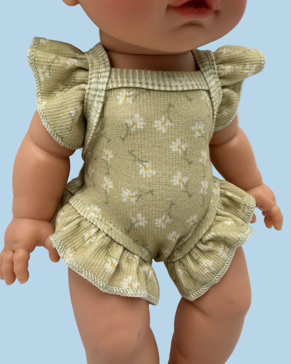 A close-up of a doll wearing a ribbed sage green romper with a small white floral print, ruffled shoulder straps, and ruffled leg openings.